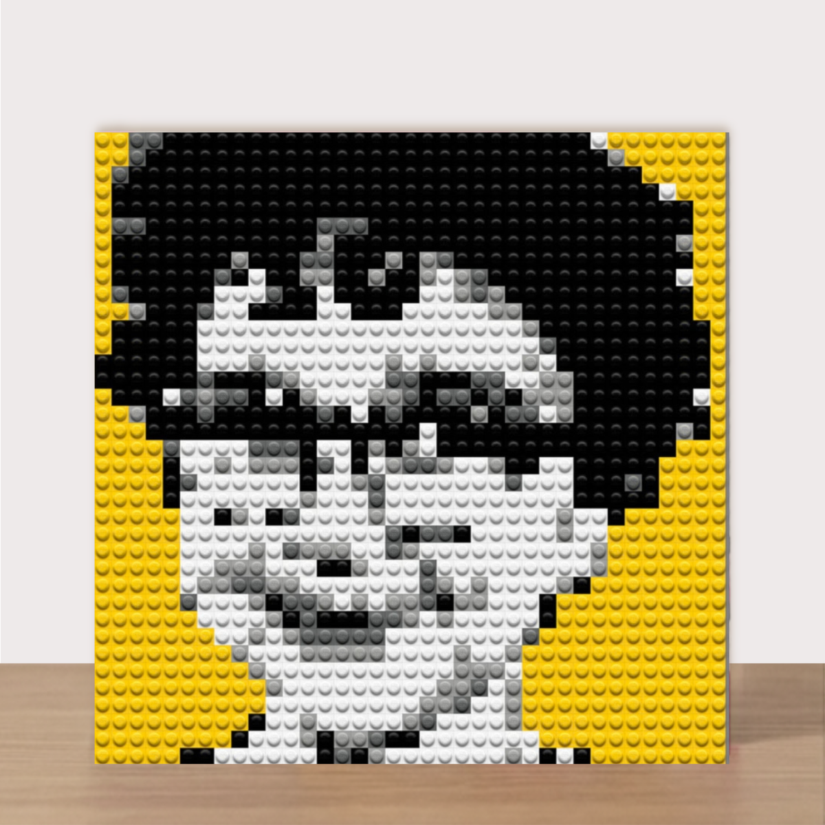 A square custom portrait mosaic built from interlocking toy bricks, featuring a black, white, and gray pixelated face on a yellow and black background, displayed against a light gray wall and wooden floor.