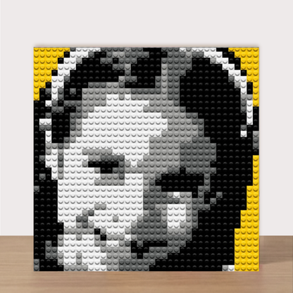 Finished Bricky Cam Pix girl hero pixel art. A square brick-built portrait artwork of Cam Pix girl hero face constructed from black, white, and gray toy studs, with yellow side accents, displayed against a light wall above a wood surface.